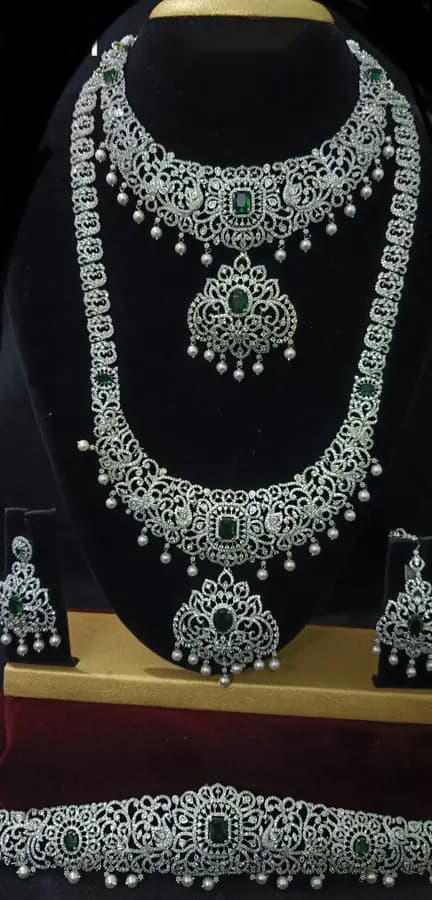 American diamond bridal jewellery set on a display stand — short necklace, long haram, jhumka and oddiyanam, with green centre stones