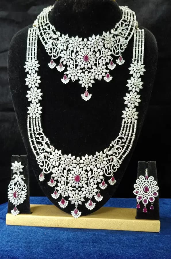 American diamond bridal jewellery set on a display stand — choker, long haram, jhumka and oddiyanam, with red stones
