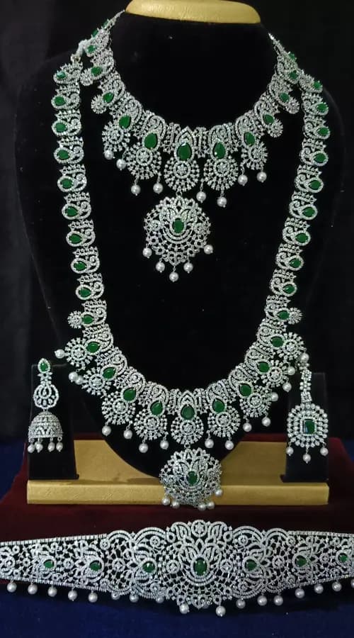 American diamond bridal jewellery set on a display stand — choker, long haram, jhumka and oddiyanam, with green stones