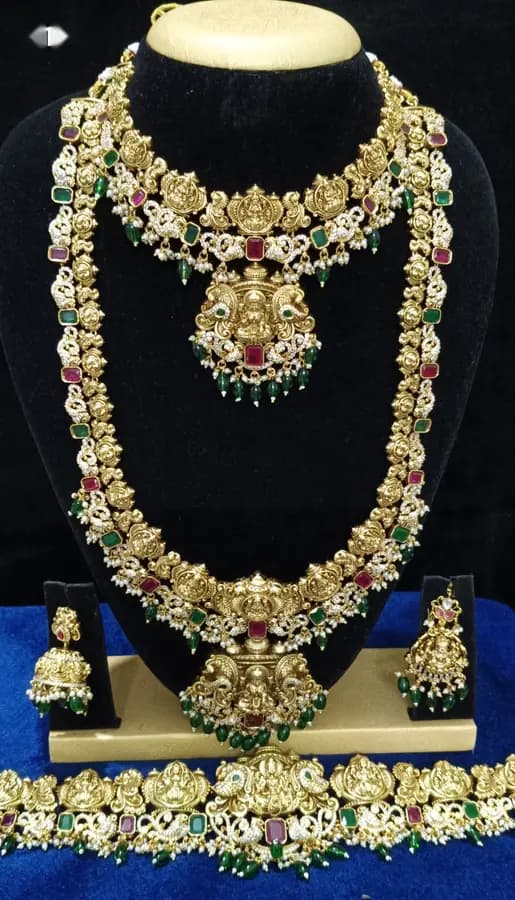 Antique gold temple jewellery bridal set on a display stand — layered necklace and haram, jhumka and oddiyanam, with multicoloured stones