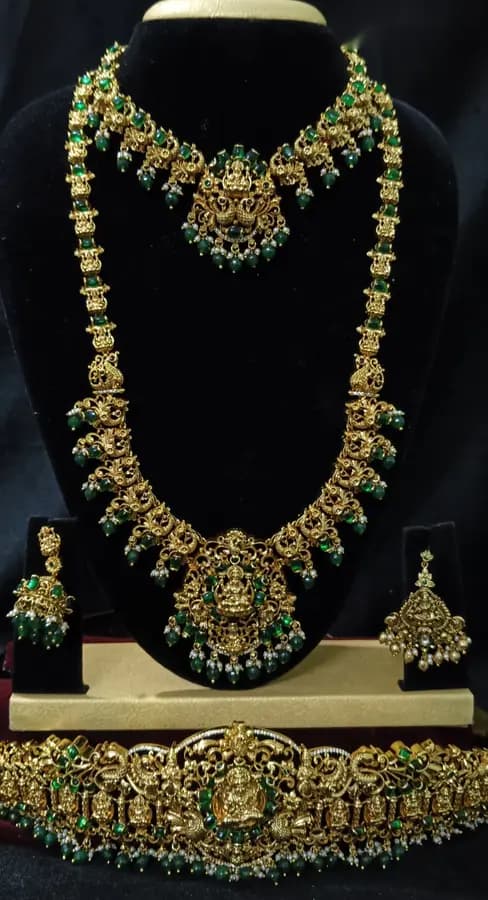 Antique gold temple jewellery bridal set on a display stand — short necklace, long haram, jhumka and oddiyanam, with green beads
