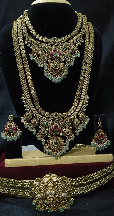 Antique gold temple jewellery bridal set on a display stand — choker, necklace, long haram and oddiyanam, with red and green stones