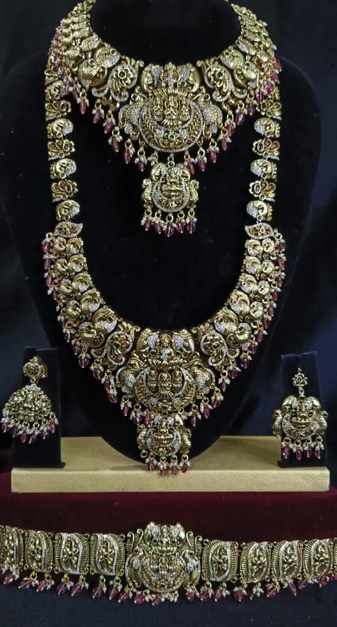 Antique gold temple jewellery bridal set on a display stand — short necklace, long haram, jhumka and oddiyanam, with red stones