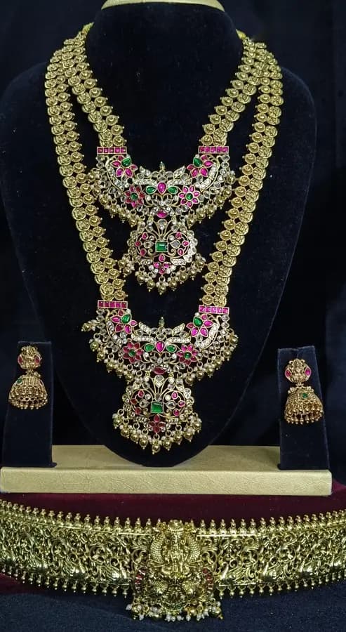 Antique gold temple jewellery bridal set on a display stand — layered necklace and haram, jhumka and a wide oddiyanam, with pink and red stones