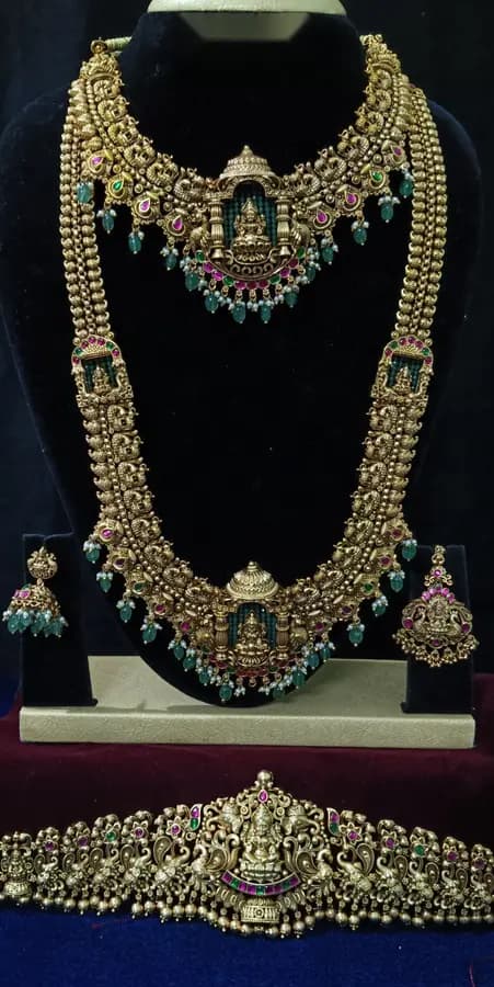 Antique gold temple jewellery bridal set on a display stand — choker, long haram, jhumka and oddiyanam, with green stones