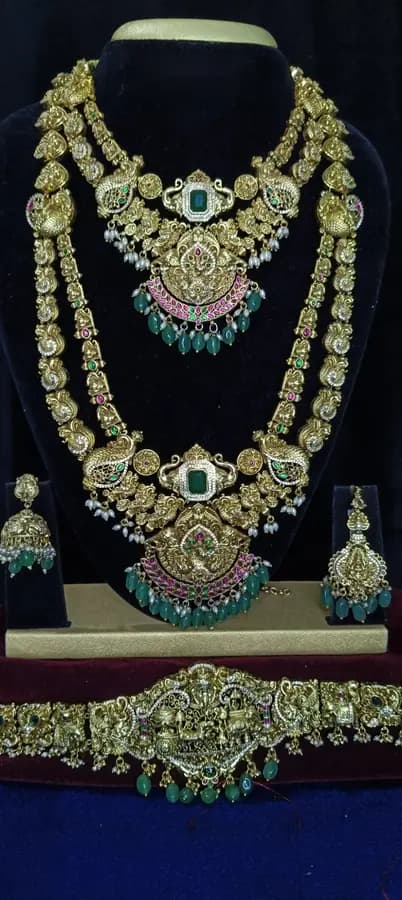 Antique gold temple jewellery bridal set on a display stand — short necklace, long haram, jhumka and oddiyanam, with green drops