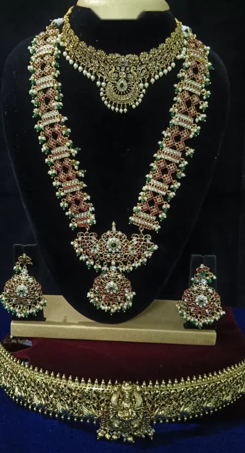 Antique gold temple jewellery bridal set on a display stand — short necklace, long haram, jhumka and oddiyanam, with red stones