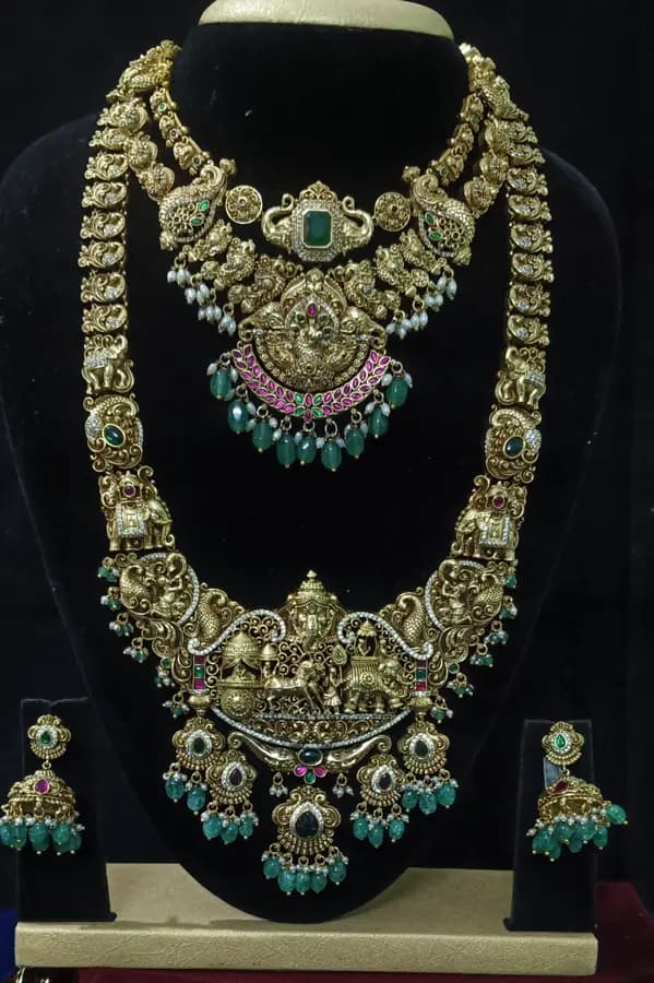 Antique gold temple jewellery bridal set on a display stand — layered necklace and haram with a large Lakshmi pendant and jhumka, with green bead drops